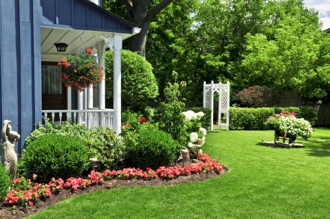 A Back Yard With Covered Porch And Hanging Flower Baskets With Manicured Lawn, Flower Beds, Shrubs, Statues, And A Trellis.