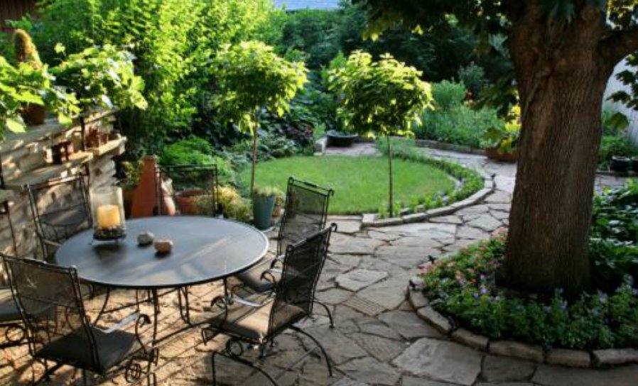 Outdoor Stone Patio Seating Area With Table And Chair, Tree With Garden Bed Surrounding It, And Manicured Lawn