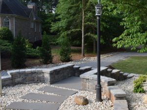 Short Rock Wall And Lamp Post With Pebble Landscaping And Stone Walkway. Green Shrubbery And Manicured Lawn In The Background.